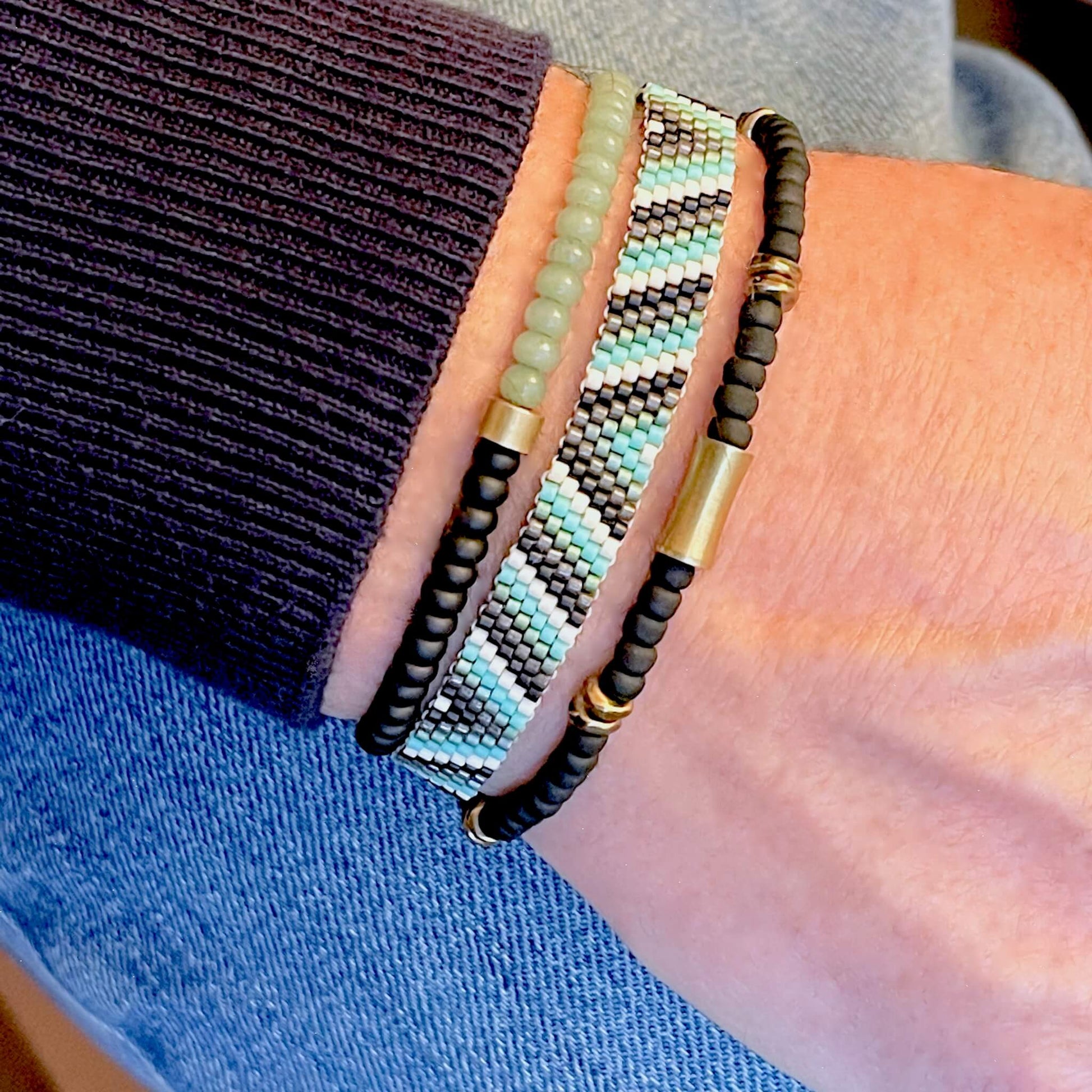 Men's beaded bracelet stack with a woven black, aqua and white seed bead band and two black and green stretch bracelets with raw brass accents.