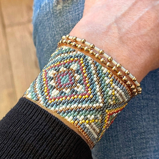 Hand woven peyote beaded bracelet in muted yellow, sage, and gray diamond pattern, stacked with gold ball beaded stretch bracelets.