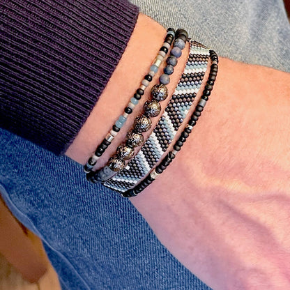 Men’s handwoven blue & black beaded bracelet stacked with stretch bracelets of gunmetal lava, blue dumortierite, & seed beads
