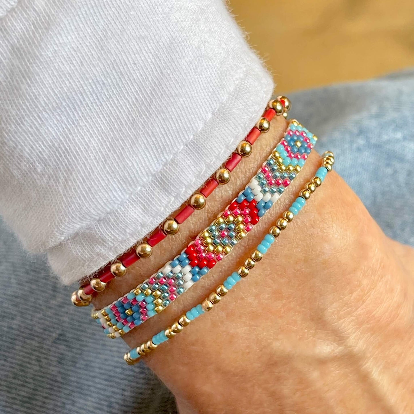 Red and blue woven bracelet stacked with 2 gold ball stretch bracelets wtih glass seed beads.